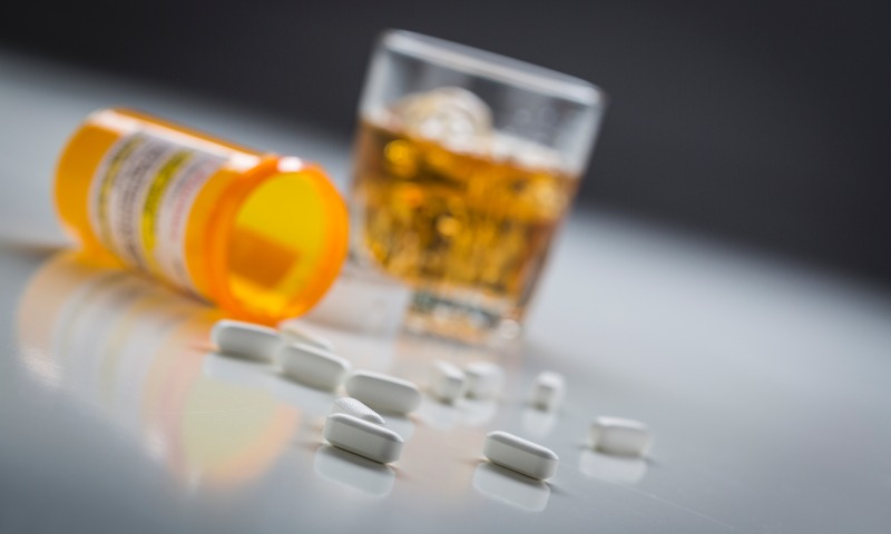 Prescription pills spilled from bottle near glass of alcohol.