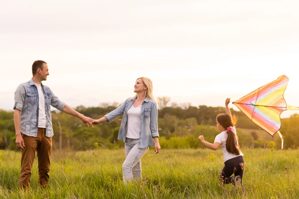Family flying kite together in meadow at sunset.